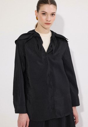 Kobieta o brązowych włosach związanych do tyłu, ubrana w czarną koszulę na guziki z długim rękawem i dużymi kokardami przy kołnierzu, na beżowym topie, na białym tle.