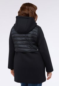 Schwarzer Mantel mit Kapuze, ausgestattet mit einem gesteppten, gepolsterten Rückenpaneel. Glatte Textur, mittellanges Design und Druckknopf-Details an der Taille.