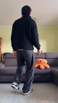 Person i svart tröja och jeans som går mot en grå soffa med en orange bläckfiskplush i ett vardagsrum.