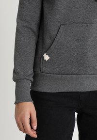 Persona che indossa un felpa grigia scura con una tasca strappata e pantaloni neri, braccio sinistro rilassato lungo il corpo.