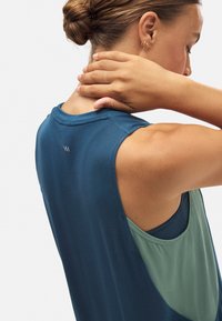 Mujer con el cabello recogido en un moño, vistiendo ropa atlética sin mangas en azul y verde, tocándose la parte posterior del cuello con la mano derecha.