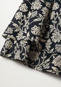 Tejido negro con un patrón de flores y viñas en color blanco roto, presentado en pliegues superpuestos sobre un fondo blanco.