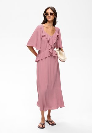 Femme portant une robe midi mauve à volants, des lunettes de soleil, des sandales noires et tenant un sac blanc plié sur un fond blanc uni.