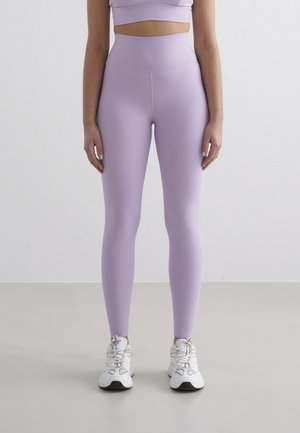 Personne portant des leggings lavande taille haute, un top court assorti et des chaussures de sport blanches, debout sur un sol uni contre un mur blanc.