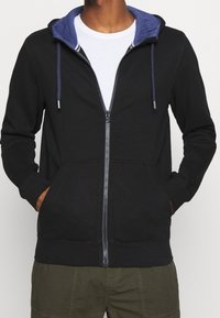 Sweat à capuche noir zippé avec capuche intérieure bleue, doté de cordons, poches frontales et poignets côtelés. Tissu doux avec texture lisse.