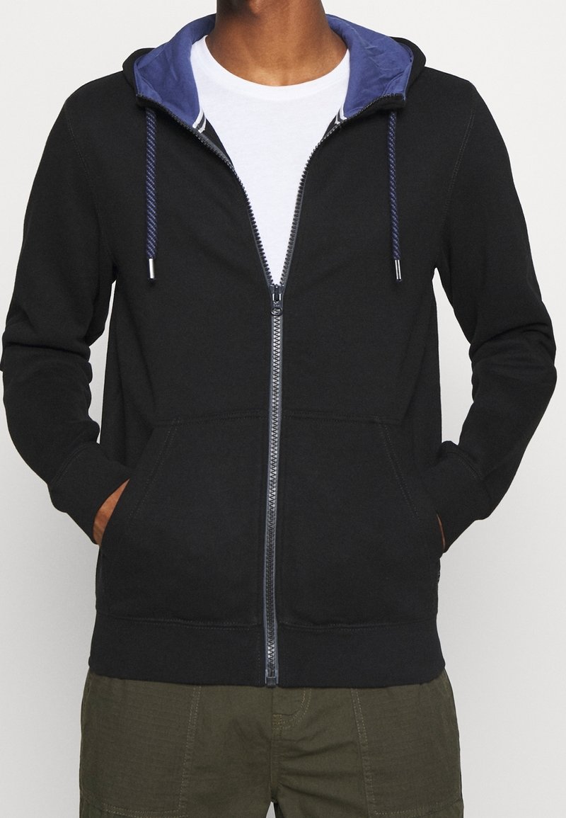 Sweat à capuche noir zippé avec capuche intérieure bleue, doté de cordons, poches frontales et poignets côtelés. Tissu doux avec texture lisse.