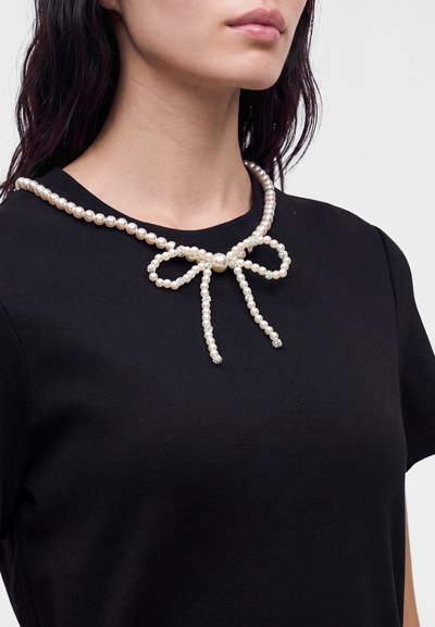 Femme portant un haut noir avec un collier de perles en forme de nœud autour du cou.