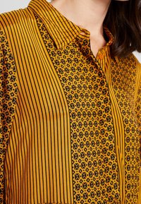 Hemd mit Knopfleiste, das ein gelb-schwarz gestreiftes und gemustertes Design aufweist, mit einem Kragen und einem strukturierten Stoff. Auffälliges orangefarbenes Knopfdetail.