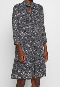 Floral print dress in navy with three-quarter sleeves, collar, keyhole opening, and pleated skirt. Features small blue, white, and brown flowers.