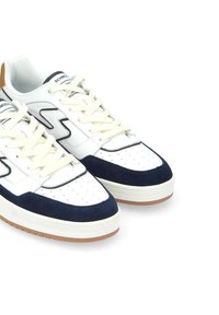 Weiße Sneakers mit marineblauen Wildlederelementen, perforiertem Zehenbereich und cremefarbenen Schnürsenkeln. Sie verfügen über eine gepolsterte weiße Sohle und eine braune Gummilaufsohle.