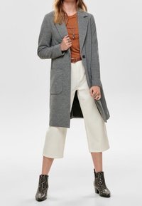 Donna che indossa un cappotto lungo grigio, una maglia marrone, pantaloni bianchi a gamba larga e corti, collane a strati e stivaletti con stampa a serpente, in piedi con le gambe divaricate.