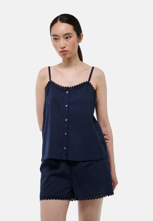 Jeune femme portant un haut sans manches bleu marine à boutons et un short assorti avec bordure en dentelle sur fond blanc.