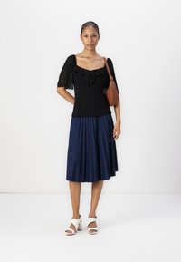 Schwarzes Oberteil mit Rüschendetails und transparenten Ärmeln, kombiniert mit einem marineblauen plissierten Rock und weißen offenen Schuhen. Inklusive einer braunen Umhängetasche.