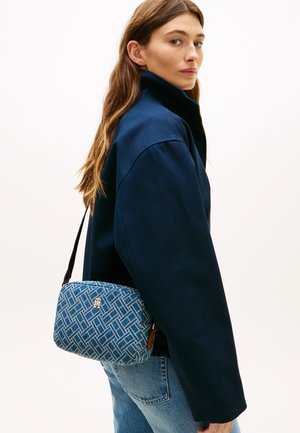 Woman with long hair wearing a navy jacket and jeans carries a blue patterned crossbody bag with a gold logo on a white background.