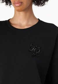 Sweatshirt en coton noir avec un embellissement en perles noires texturées en forme de cursive sur la poitrine, avec un col rond classique.