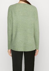 Pull vert côtelé à manches longues avec un col arrondi, présentant une coupe décontractée et une texture douce, associé à un pantalon noir texturé.