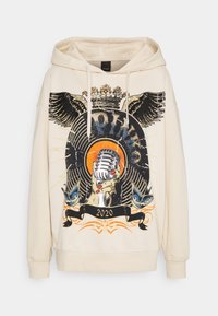 Sudadera con capucha color crema con un diseño gráfico de un micrófono, pájaros y texto. Detalles incluyen alas negras y colores vibrantes. Inscripción 2020.