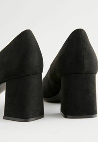 Zapatos de tacón cuadrado de ante negro con punta afilada. Presentan una superficie texturizada y un tacón grueso y cuadrado para mayor estabilidad.