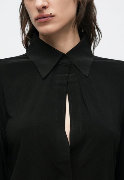 Juoda palaidinė su smailia apykakle ir vertikaliu kloste priekyje. Lygi tekstūra su švelniu blizgesiu. Minimalistinis dizainas.