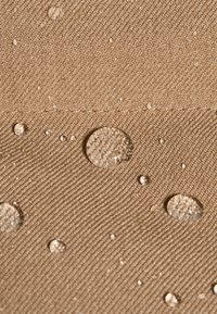 Tessuto beige con una superficie texturizzata, che presenta numerose gocce d'acqua di diverse dimensioni appoggiate su di esso. Alcune gocce sono chiare, altre leggermente sfocate.