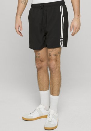 Mannlige ben iført svarte shorts med hvite striper og teksten «UNFAIR», hvite sokker og hvite joggesko med gummisåler, står på en grå bakgrunn.