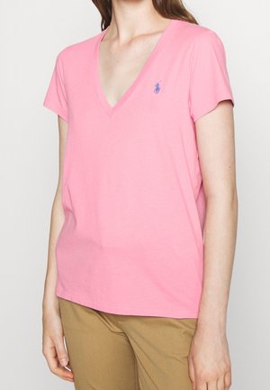 Kobieta ubrana w różowy T-shirt z krótkim rękawem i dekoltem w serek z małym niebieskim logo polo oraz beżowe spodnie, pokazana od szyi do bioder.