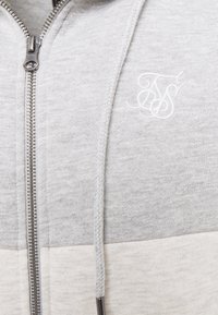 Primo piano di una felpa grigia chiaro con zip, dotata di cordino intrecciato e logo bianco ricamato sul petto.