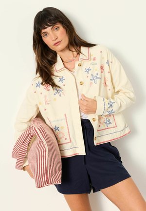 Frau mit langen braunen Haaren, die eine cremefarbene Jacke mit Seestern- und Korallenstickerei, marineblaue Shorts trägt und ein rotgestreiftes Kissen hält.