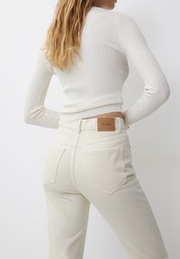 Un jean taille haute beige clair avec une texture lisse et deux poches arrière, arborant une patte en cuir marron sur la ceinture.