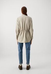 Light beige shirt with elastic waist, long sleeves, and buttoned cuffs. Paired with medium wash jeans and brown shoes, simple design.