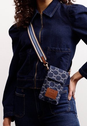Frau trägt eine marineblaue Jeansjacke und Jeans mit einer gemusterten Umhängetasche für das Telefon, versehen mit einem braunen Leder-Logo-Patch und einem gestreiften Riemen.