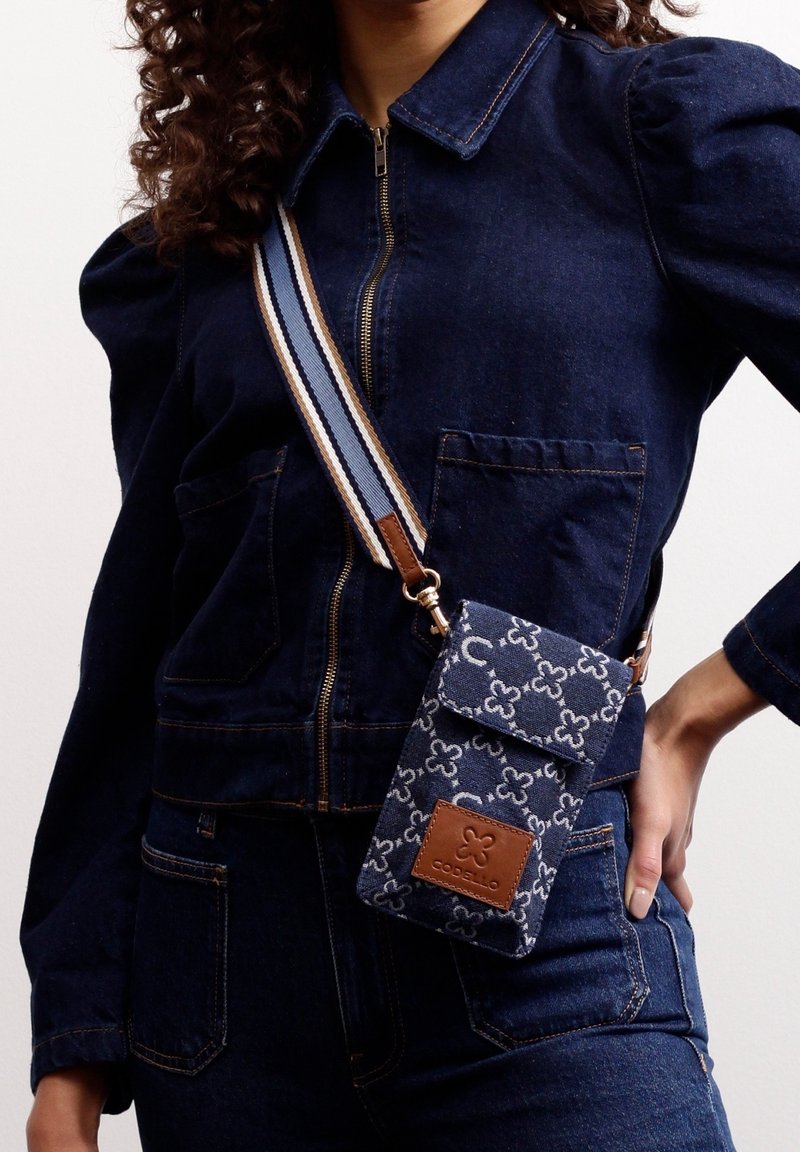Femme portant une veste en jean marine et un jean, avec un sac téléphone bandoulière à motifs, comprenant une pièce en cuir marron avec logo et une bandoulière rayée.