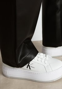 Sneakers bianchi con suola testurizzata, caratterizzati da una punta arrotondata e branding laterale. Indossati con pantaloni neri a gamba affusolata in pelle sintetica.