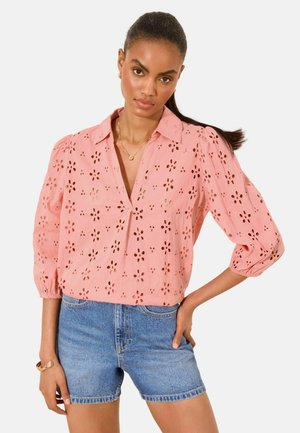 Žena v ružovej kvetinovej eyelet blúzke s 3/4 puff rukávmi a riflových kraťasoch pózujúca s rukou v páse na bielom pozadí.