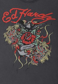 Camiseta negra con arte colorido estilo tatuaje que presenta un león, una rosa y un águila, acentuada por la audaz letra roja de "Ed Hardy."