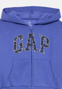 Felpa blu per bambini con zip, cappuccio, tasche frontali e testo "GAP" in un motivo floreale nero contornato di bianco.