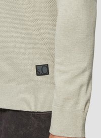 Teksturowany jasnobeżowy sweter z prążkowanymi mankietami i dołem, z czarną kwadratową metką logo na dolnym lewym boku.