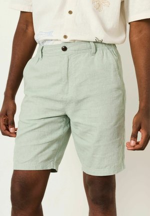 Mann trägt hellgrüne Leinen-Shorts mit Knopfverschluss und Gürtelschlaufen, kombiniert mit einem weißen Kurzarmhemd mit Blumenstickerei.