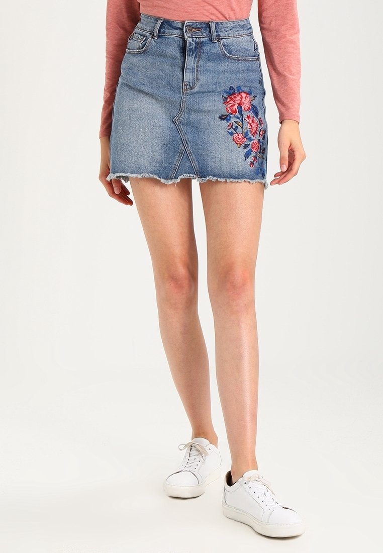 Jupe en denim avec un ourlet effiloché, ornée d'une broderie florale sur le côté gauche. Design classique avec cinq poches et fermeture à boutons sur le devant.
