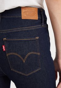 Tmavě modré džíny z denimu s koženou nášivkou označenou "Levi Strauss & Co." na zadní straně. Obsahují jasně žluté švy a červenou nášivku.