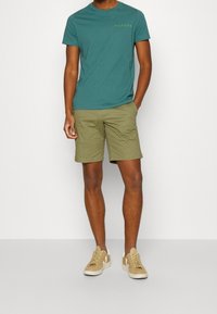 Mand iført en teal kortærmet T-shirt med "Hilfiger" tekst, olivenfarvede shorts og beige sneakers med hvide såler og logo.