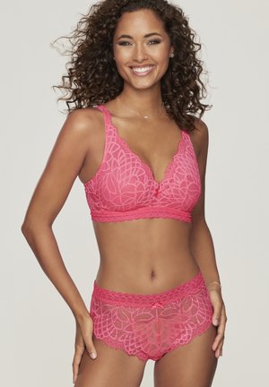Lächelnde Frau mit lockigem Haar, die ein passendes rosa Spitzen-BH und hoch taillierte Höschen mit Blumenmuster trägt und vor einem hellen Hintergrund posiert.