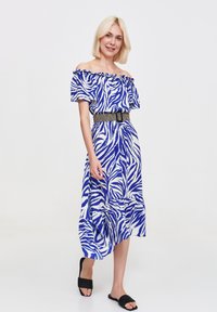 Kobieta z krótkimi blond włosami uśmiecha się, ubrana w niebiesko-białą sukienkę w zebrę na jedno ramię i czarne sandały na płaskiej podeszwie, stojąc na jednolitym tle.