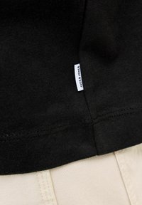 Černé bavlněné tričko s hladkou texturou, s malou bílou štítkem s nápisem „JACK & JONES“ přišitým na lemu, doplněné světle zbarvenými kalhotami.