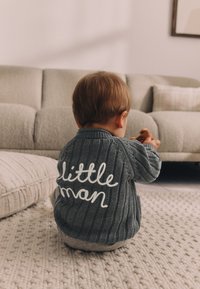 Pull gris en tricot avec texture côtelée et texte blanc "petit homme" au dos, porté par un enfant assis sur un tapis texturé.