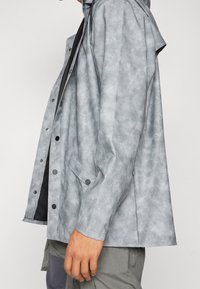 Jasnoszara, teksturowana kurtka z kapturem, z napami, kieszenią z przodu i luźnym fasonem. Wewnątrz podszewka jest czarna.