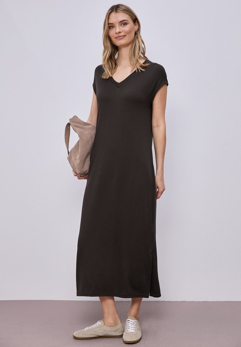 Vrouw die een zwarte midi-jurk met V-hals en kapmouwen draagt, een beige suède tas vasthoudt en beige sneakers draagt, staand tegen een effen achtergrond.