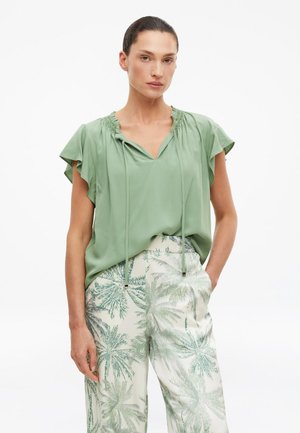 Donna che indossa una camicetta verde chiaro con maniche svolazzanti e scollo con coulisse, abbinata a pantaloni bianchi con stampe di palme verdi.