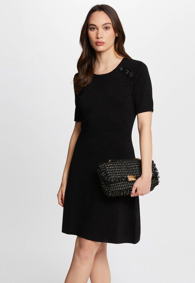 Zwarte jurk met korte mouwen en een ronde halslijn, versierd met schouderdetails, gecombineerd met een gestructureerde zwart-wit clutch.