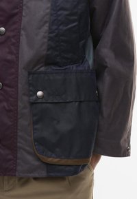 Viaszos anyagú dzseki sötét tengerészkék és burgundi árnyalatokban. Gombos zsebekkel és diszkrét logódíszítéssel az elején.